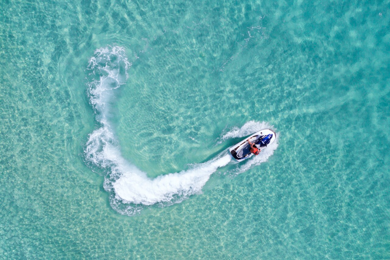 Aerial view of a tourist enjoying a high-speed jet ski ride creating a wake in clear turquoise tropical ocean waters.