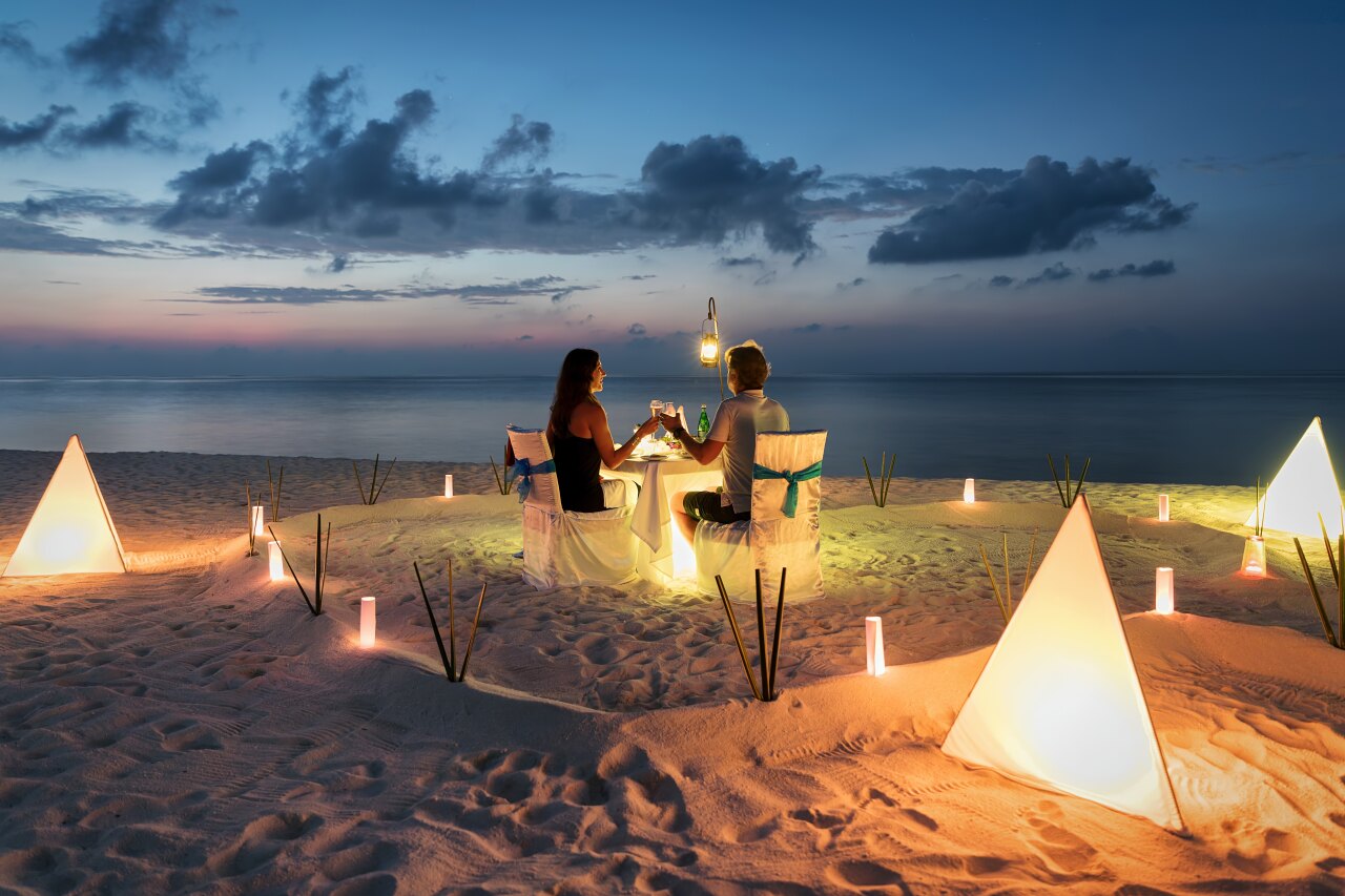 Una coppia si gode una cena romantica privata su una spiaggia di sabbia bianca al tramonto con lanterne piramidali.