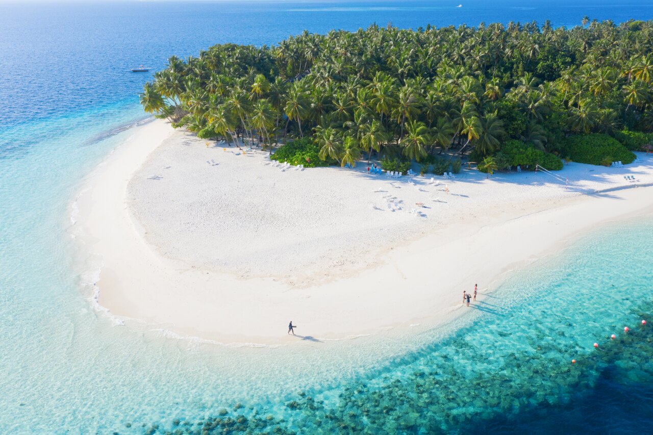 Aerial drone shot of a white sandbank surrounded by turquoise water and lush green palm trees at a luxury resort.