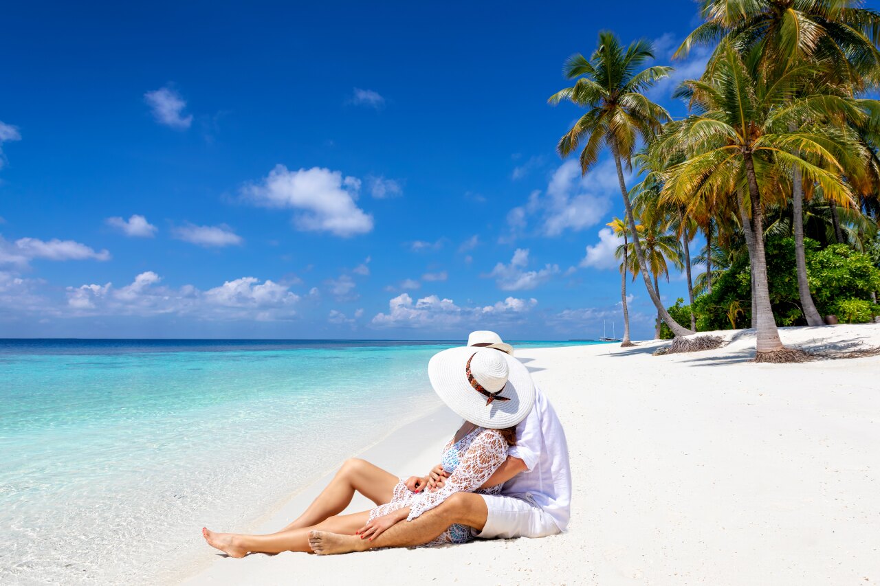 A romantic couple wearing sun hats sitting on a beautiful white sand beach with turquoise water and palm trees.