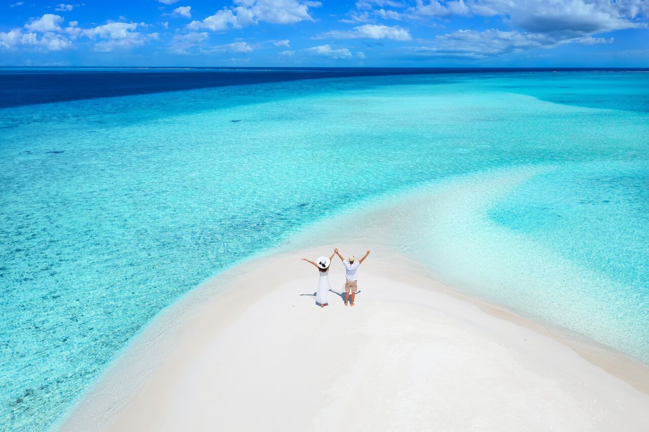 Aerial view of a happy couple standing on a white sandbank surrounded by turquoise water under a clear blue sky