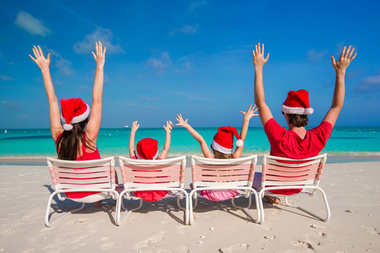 Back view of a happy family of four wearing Santa hats sitting on beach chairs on a sunny tropical sandy beach.