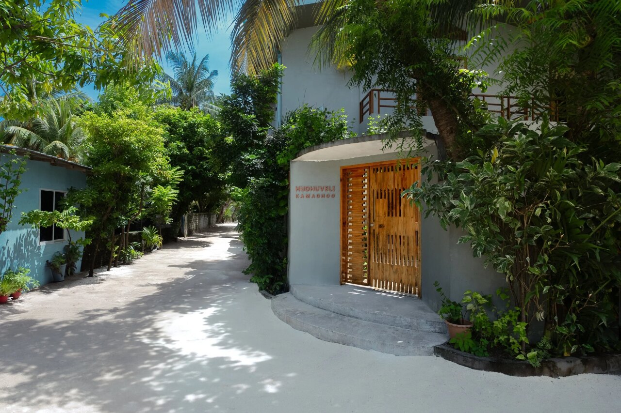 Il cancello in legno e l'ingresso della guesthouse Hudhuveli Kamadhoo circondati da palme tropicali e vegetazione.