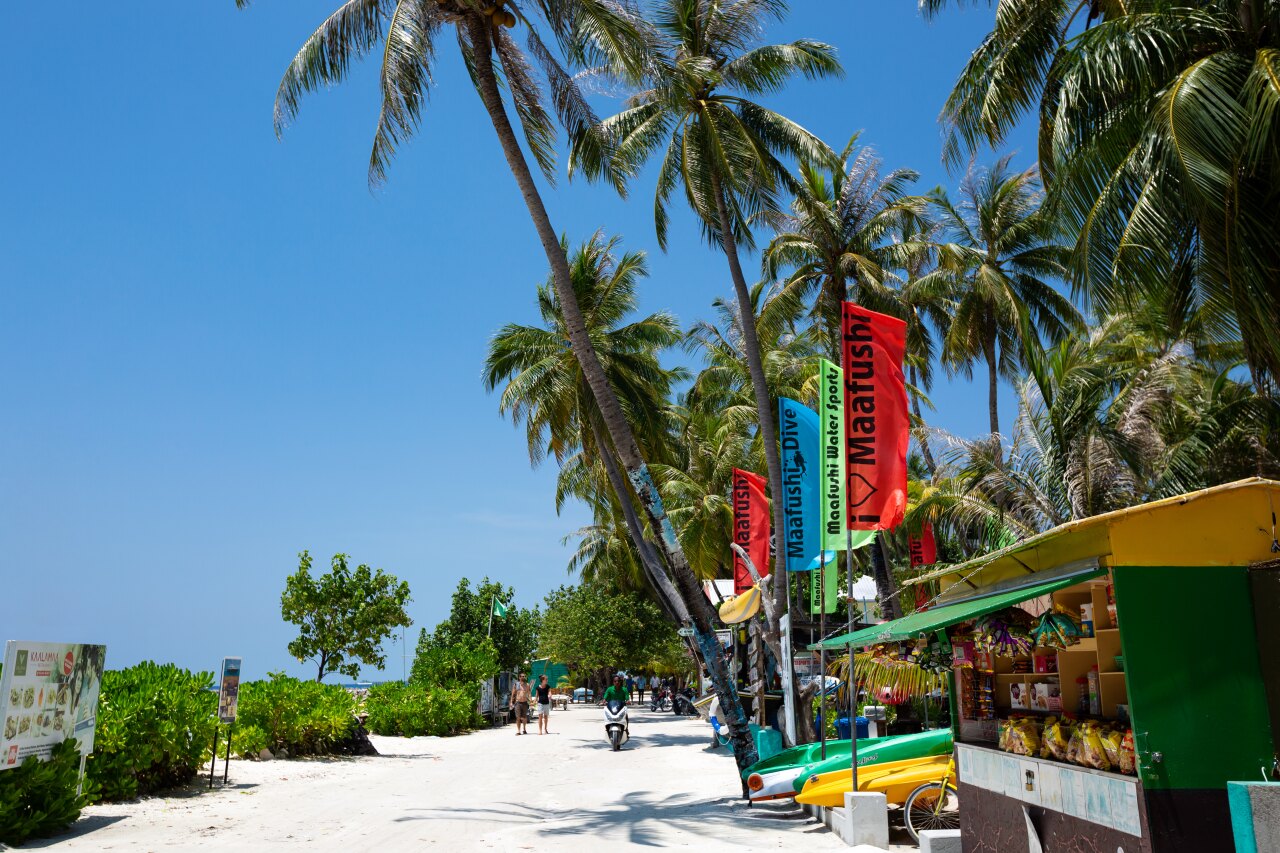 Sunny sandy street with palm trees, water sports flags, and a snack shop in Maafushi, Kaafu Atoll, Maldives