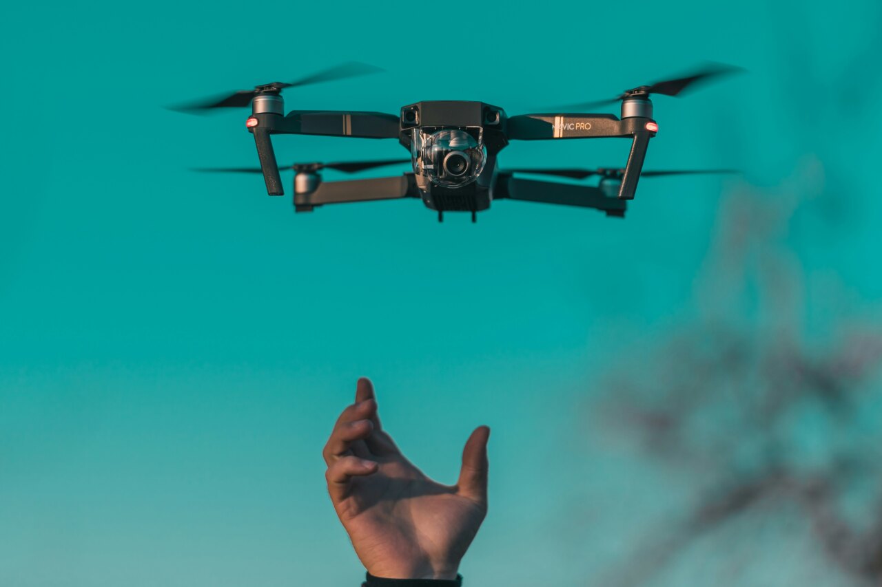 A black DJI Mavic Pro drone hovering in the air as a hand reaches up to catch it against a bright cyan sky background