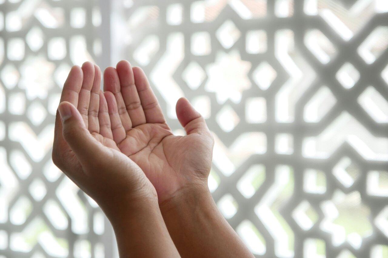 Primo piano delle mani di una persona alzate in preghiera, su uno sfondo sfocato con un tradizionale schermo islamico.