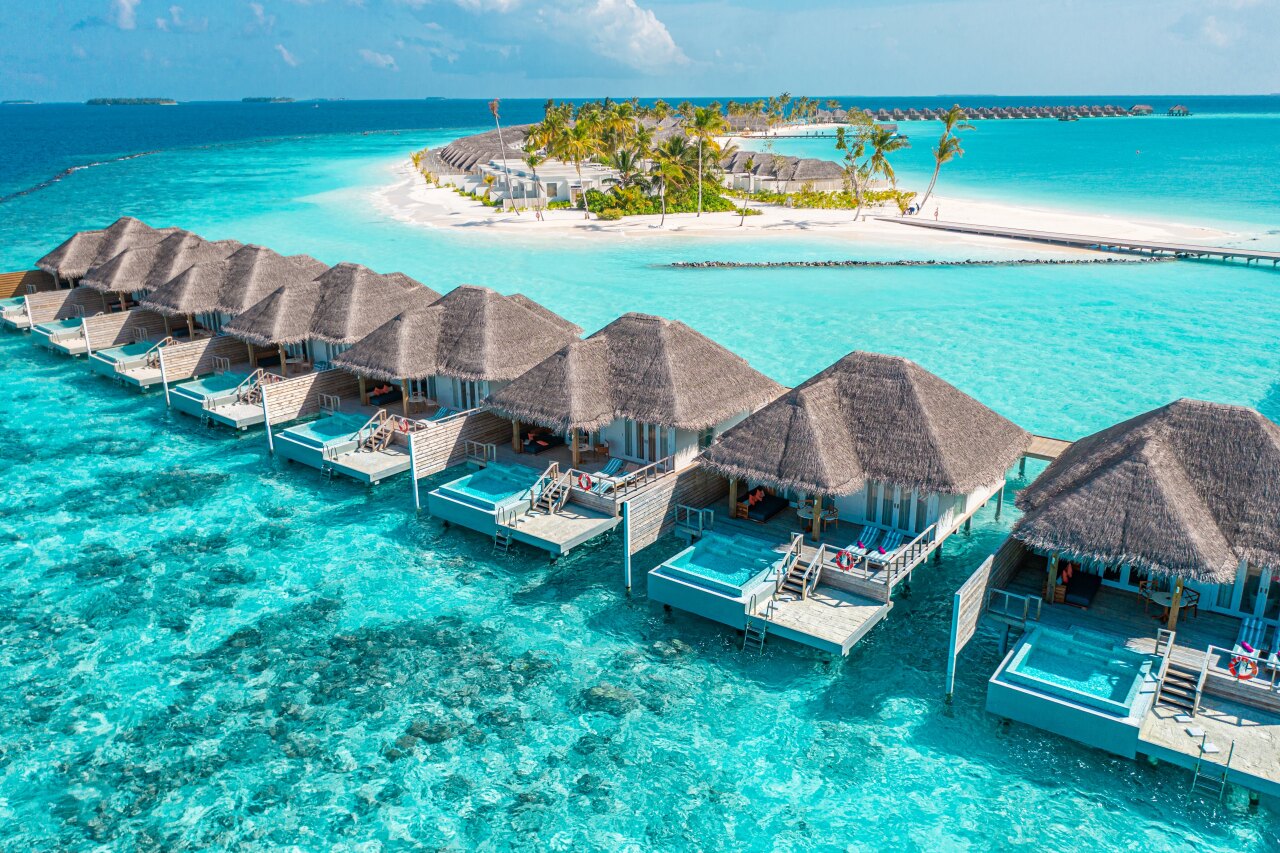 Aerial drone view of a row of wooden overwater villas with private infinity pools sitting above turquoise coral reef