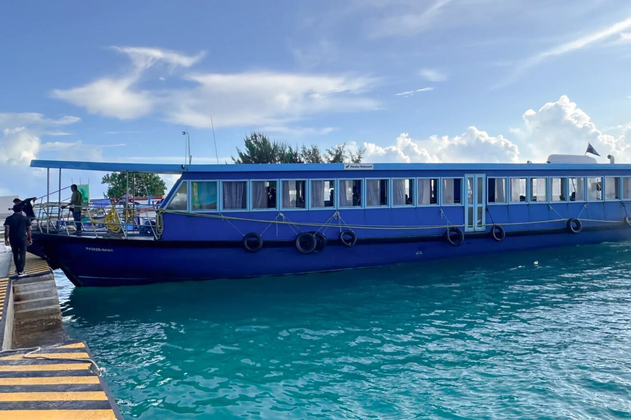 A blue public ferry boat tied to a concrete pier surrounded by clear turquoise water under a blue cloudy sky.