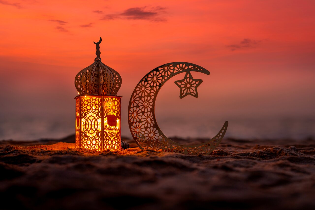 Lanterna decorativa di Ramadan e ornamento a forma di mezzaluna sulla spiaggia sabbiosa durante un tramonto arancione.