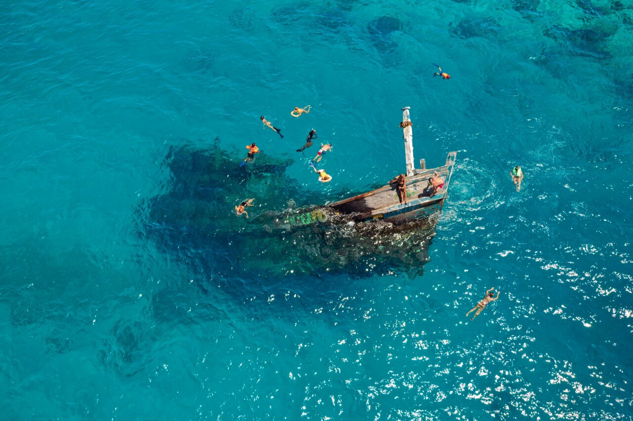 Aerial view of tourists snorkeling in clear blue water around a sunlit wooden shipwreck in the vaavu atoll in Maldives