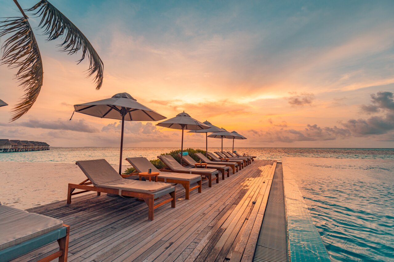Wooden deck with row of sun loungers and umbrellas beside an infinity pool during a scenic golden hour sunset.