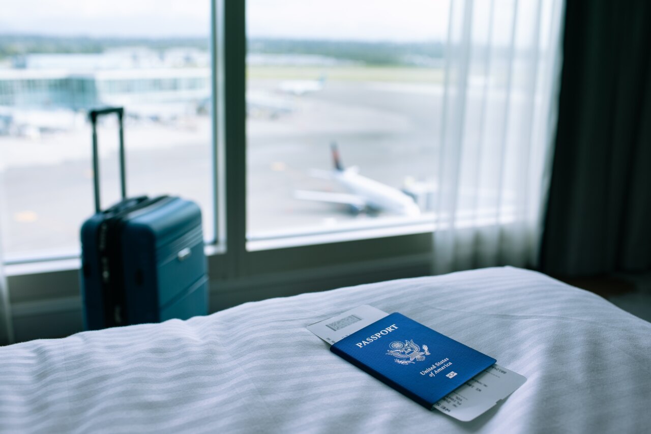 A blue United States passport with travel documents lies on a white bed sheet near a window overlooking an airport runway.
