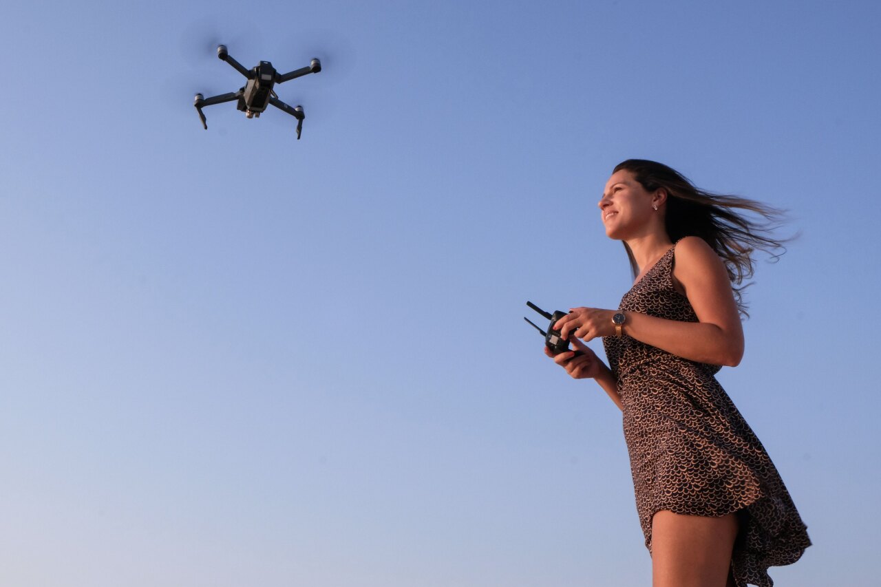 A smiling young woman in a summer dress standing outdoors while controlling a flying camera drone against clear blue sky