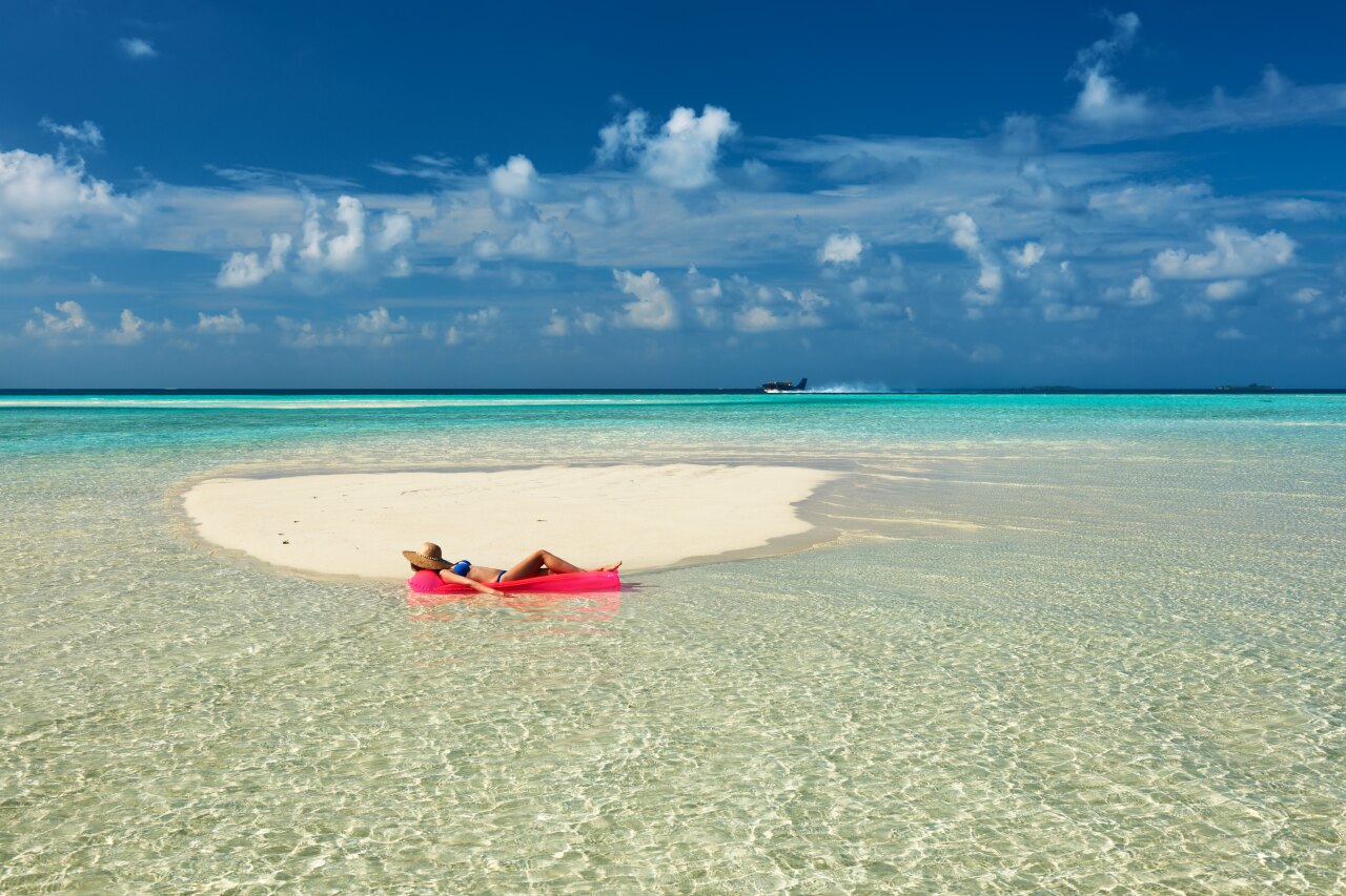 A woman wearing a straw hat rests on a bright pink inflatable mattress in the shallow crystal water of a sandbank