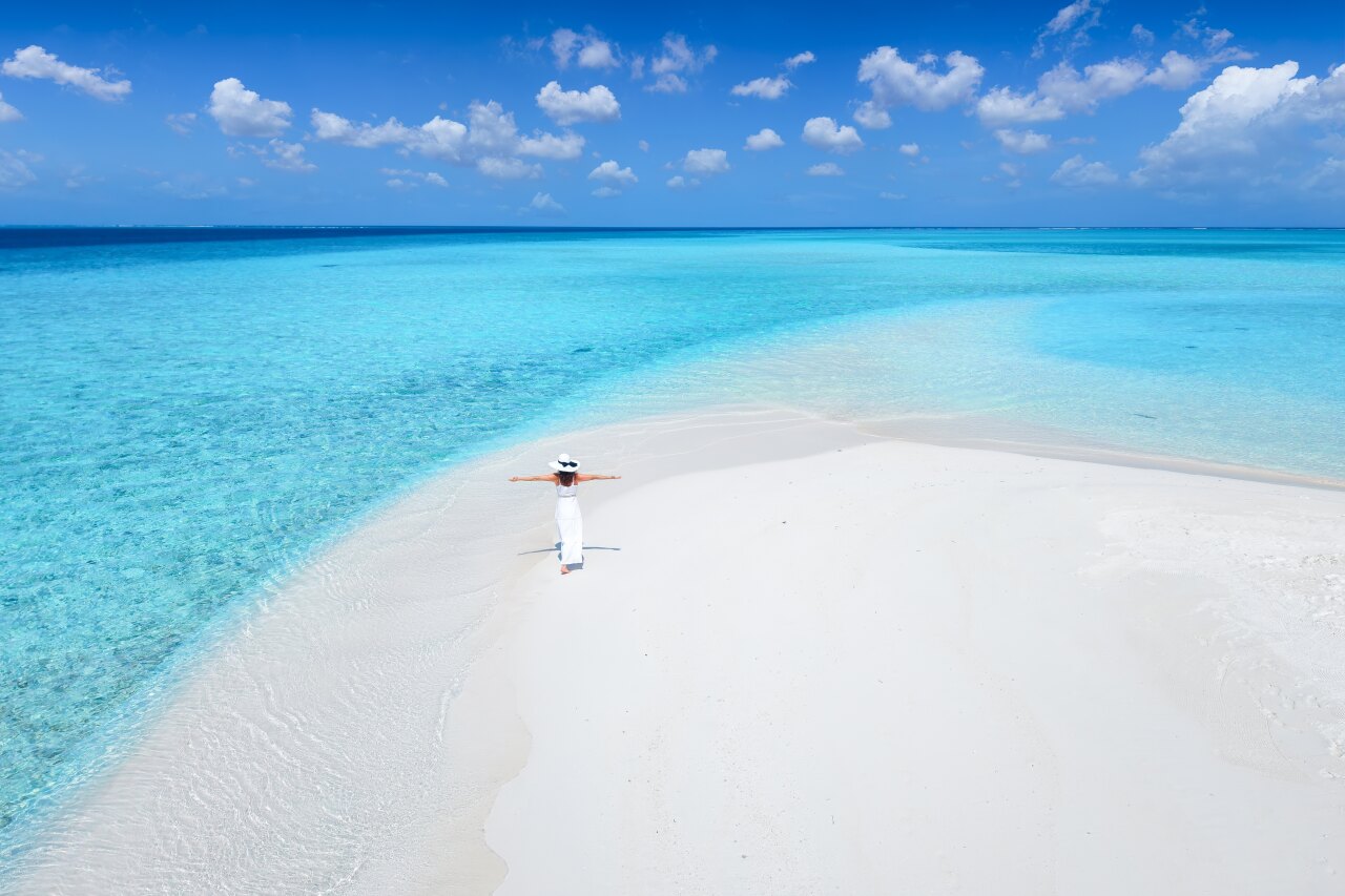 A woman in a white dress and hat standing with arms open on a pristine white sandbank surrounded by turquoise sea