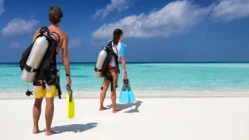 activity, people, sea - A man and woman carrying scuba tanks and fins on a pristine white sandy beach, preparing to dive in tropical water.