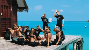 activity, people - A group of scuba divers and instructors in wetsuits sitting on a wooden jetty by the turquoise ocean in the Maldives