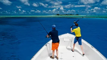 activity, sea - Two men on a boat casting their fishing rods into the vibrant blue ocean with a remote tropical island in distance