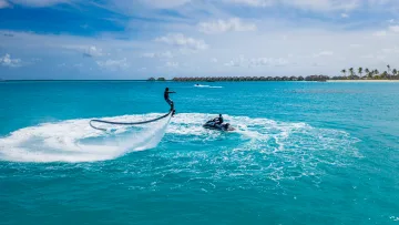 activity, resort, sea - A person soaring above the crystal blue sea on a flyboard with a jet ski nearby, surrounded by tropical resort scenery.