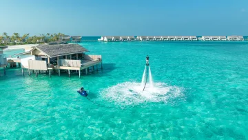 activity, resort, sea - Aerial view of a person performing flyboard stunts on turquoise water near an overwater villa at a luxury resort.