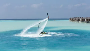activity, resort, sea - A person performing a flyboard stunt above the crystal clear turquoise tropical water near resort water villas.