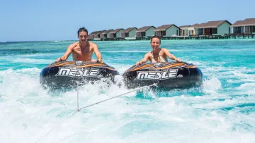 activity, people, resort, sea - A happy couple enjoys a thrilling funtube water sport ride on clear turquoise sea in front of luxury water villas.