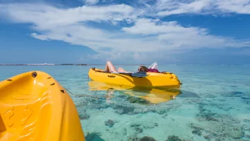 activity, reef, relax, sea - Una donna distesa su un kayak giallo in acque basse turchesi con barriera corallina visibile sotto un cielo blu