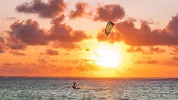activity, sky, sunset - A silhouette of a person kitesurfing across the ocean during a vibrant golden sunset with dramatic orange clouds.