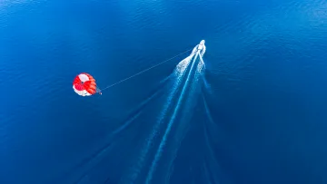 activity, drone-shot, sea - Aerial view of a vibrant red and white parachute being pulled by a speed boat over the deep blue tropical ocean.