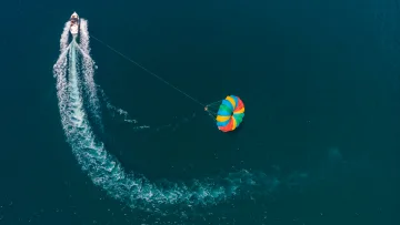 activity, drone-shot, sea - Aerial view of a colorful parasail parachute towed by a speed boat, leaving a white wake in the deep blue ocean water.