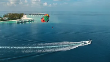 activity, drone-shot, resort, sea - Aerial view of a colorful parasail soaring above the blue ocean near a luxury resort island with overwater villas.