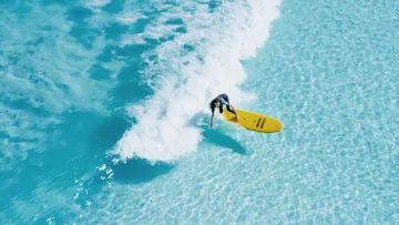 activity, drone-shot, sea - Drone shot of a surfer riding a small wave on a yellow board in crystal clear turquoise shallow tropical waters.