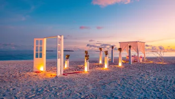 activity, beach, luxury, sunset - A romantic beach wedding ceremony setup with illuminated white doors and a canopy under a stunning sunset sky.