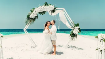 activity, beach, people - A romantic couple sharing a tender moment during their wedding ceremony under a floral arch on a pristine beach.