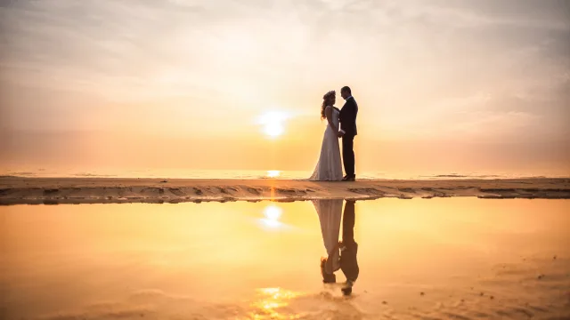 activity, beach, people, sunset - A romantic newlywed couple holding hands on a tropical island beach during a golden sunset, with their reflection visible in the wet sand.