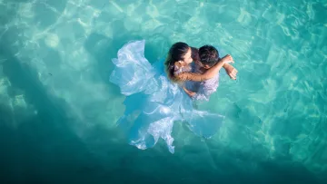 activity, people, sea - Aerial view of a newlywed couple kissing while floating in the clear turquoise ocean water in their wedding attire.