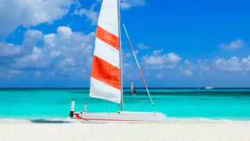 activity, beach - A white catamaran with red and white striped sails rests on a tropical sandy beach with turquoise water background.
