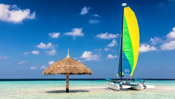 activity, beach - A colorful catamaran sits on a white sandy beach next to a wooden straw parasol in a stunning turquoise lagoon.