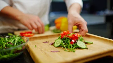 food - A chef in a white uniform carefully arranges a fresh salad with sliced cucumbers and peppers on a wooden board.