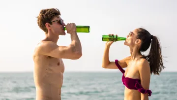 people, resort - Giovane coppia in forma con occhiali da sole che beve birre fredde in bottiglia sulla spiaggia durante la vacanza