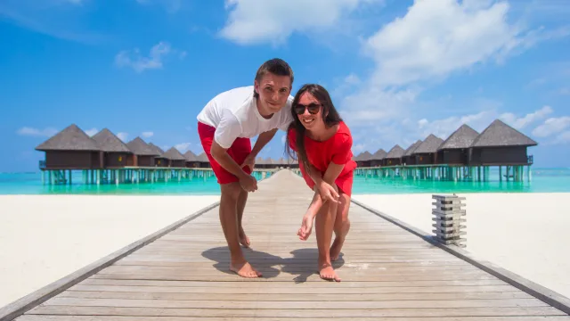 people, resort - A happy young couple leaning forward on a wooden jetty with overwater bungalow villas in the background under blue sky