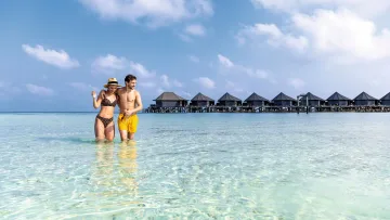 people, resort, sea - A young couple in swimwear walking through the crystal clear turquoise water of a lagoon with water villas behind.