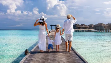 people, resort - Una famiglia di quattro persone cammina su un pontile in legno verso i bungalow sull'acqua sotto un cielo azzurro.