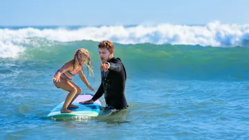 activity, people - A professional surf instructor helps a young girl balance on a colorful surfboard in the turquoise ocean waves.