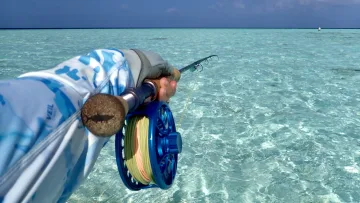 activity, sea, sky - First-person view of a person holding a fly fishing rod with a blue reel over clear, shallow turquoise tropical sea water under a sunny sky.