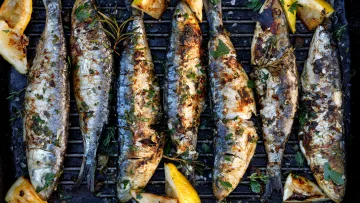 food - Top-down view of six grilled sardines on a black ribbed grill pan, garnished with herbs, coarse salt, and lemon slices.
