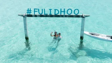 local-island, people, sea - A happy couple waving while swimming in the crystal clear turquoise water by the Fulidhoo sign in Vaavu Atoll.