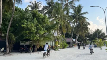 A sandy street on Maafushi Island, Kaafu Atoll, with people riding bikes and motorbikes amidst lush palm trees.