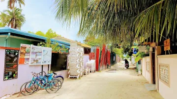 local-island - A sandy street in Maafushi local island, Kaafu Atoll with parked bicycles, stacked chairs, and a motorcycle rider.