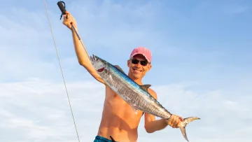 activity, people - A shirtless man with sunglasses and a pink cap smiling while holding a freshly caught wahoo fish on a fishing boat.